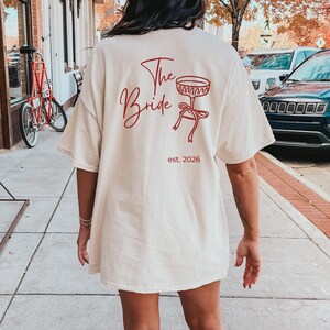 May include: Cream-colored t-shirt with the words "The Bride" in red script, along with a red graphic of a champagne glass and bow. The text "est. 2026" is below the graphic. The shirt is a casual, oversized fit.