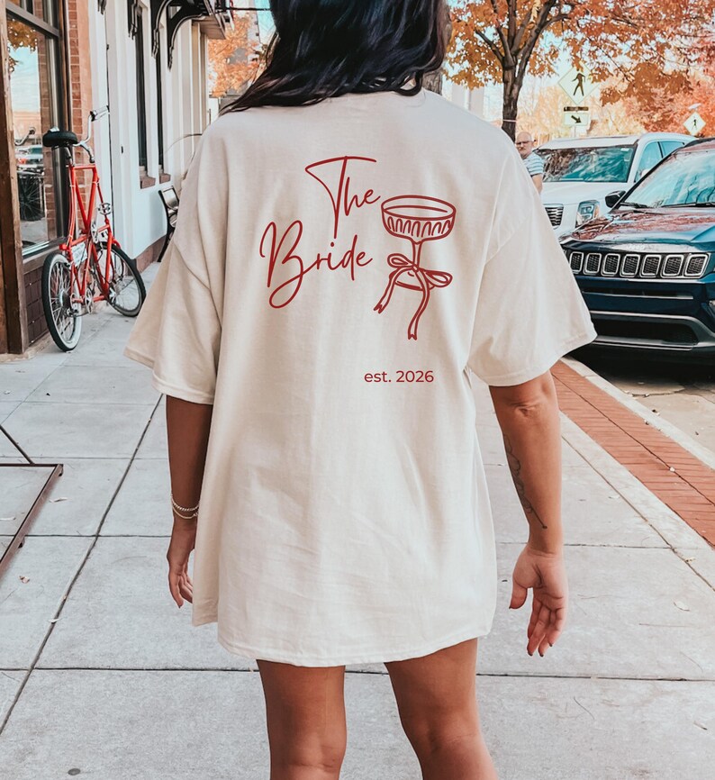 May include: Cream-colored t-shirt with the words "The Bride" in red script, along with a red graphic of a champagne glass and bow. The text "est. 2026" is below the graphic. The shirt is a casual, oversized fit.