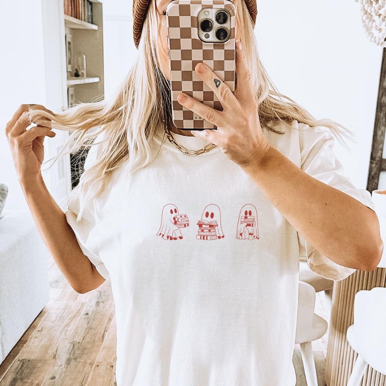 Puede incluir: Camiseta blanca con un gr&aacute;fico de tres fantasmas sosteniendo pilas de libros. Los fantasmas est&aacute;n delineados en rojo y los libros son rojos.