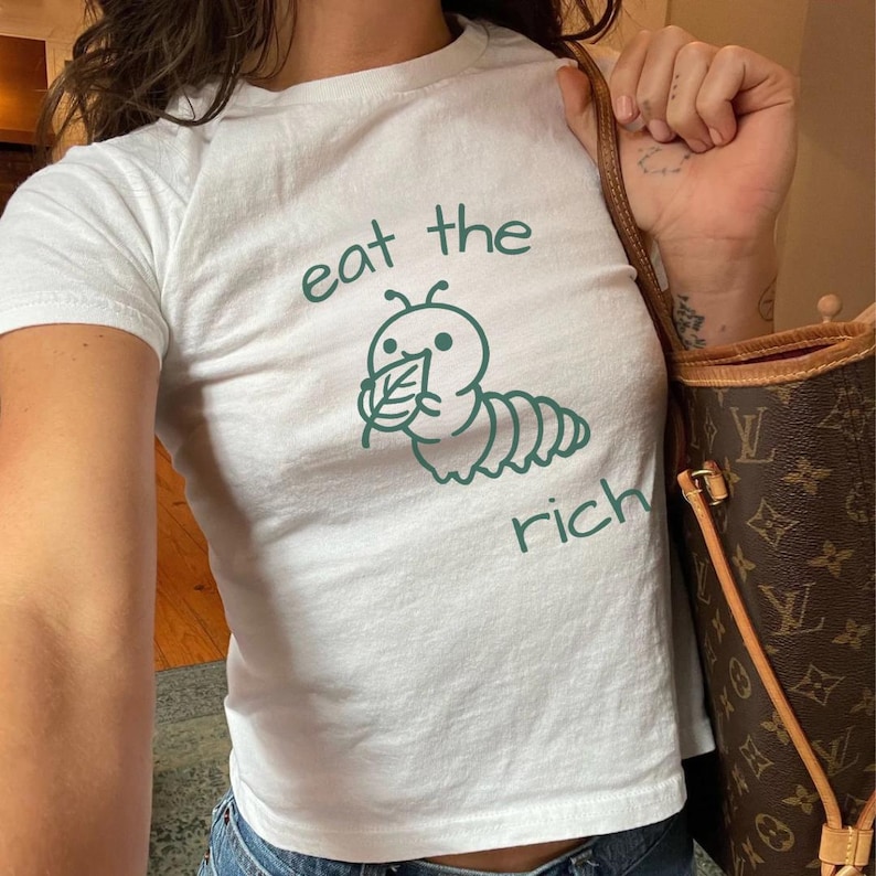 May include: A white short-sleeved t-shirt featuring a dark green graphic of a caterpillar eating a leaf. The text "eat the" is printed in dark green above the caterpillar, and the word "rich" is printed in dark green below the caterpillar.