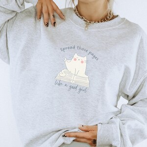 May include: A light gray crewneck sweatshirt featuring a graphic of a white cat sitting on an open book. The text above and below the cat reads, "spread those pages like a good girl." The design is printed in a dark blue color on the front center.