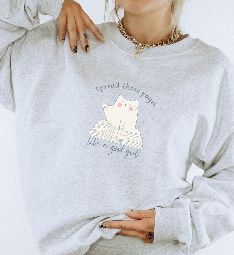 May include: A light gray crewneck sweatshirt featuring a graphic of a white cat sitting on an open book. The text above and below the cat reads, "spread those pages like a good girl." The design is printed in a dark blue color on the front center.
