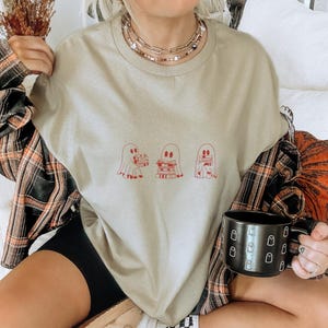 Puede incluir: Una camiseta gris claro con un gr&aacute;fico de tres fantasmas sosteniendo pilas de libros. Los fantasmas est&aacute;n delineados en rojo y los libros est&aacute;n rellenos de rojo. La camiseta la lleva puesta una persona sentada en un sof&aacute;.