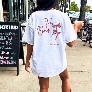 May include: White t-shirt with the text "The Bride" in red script, along with a champagne glass graphic and the date "est. 2026". The shirt is a casual, oversized fit.