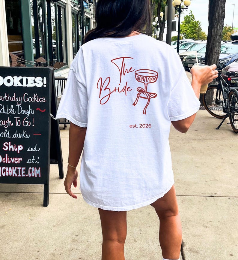 May include: White t-shirt with the text "The Bride" in red script, along with a champagne glass graphic and the date "est. 2026". The shirt is a casual, oversized fit.