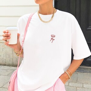 May include: White t-shirt with a small red stool graphic. The shirt is paired with pink and white striped pants and a pink shoulder bag. A gold chain necklace and bracelets complete the look. A white paper cup is held in the hand.