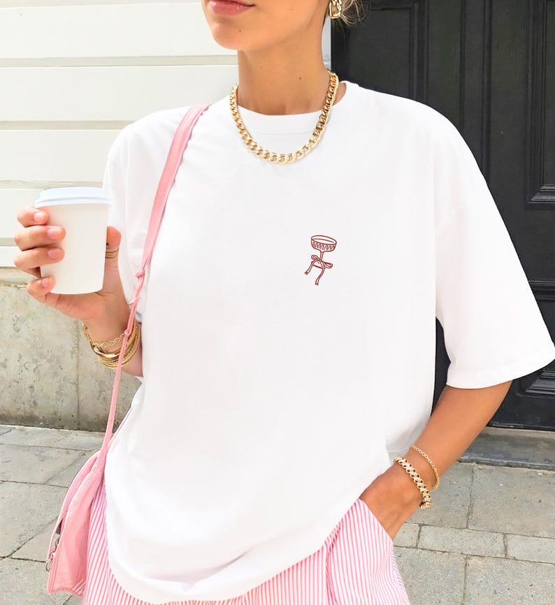 May include: White t-shirt with a small red stool graphic. The shirt is paired with pink and white striped pants and a pink shoulder bag. A gold chain necklace and bracelets complete the look. A white paper cup is held in the hand.