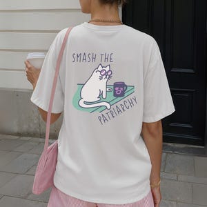 May include: Cream-colored t-shirt with a graphic on the back. The graphic features a white cat wearing sunglasses, a coffee mug, and the text "SMASH THE PATRIARCHY". The cat is sitting on a green surface.