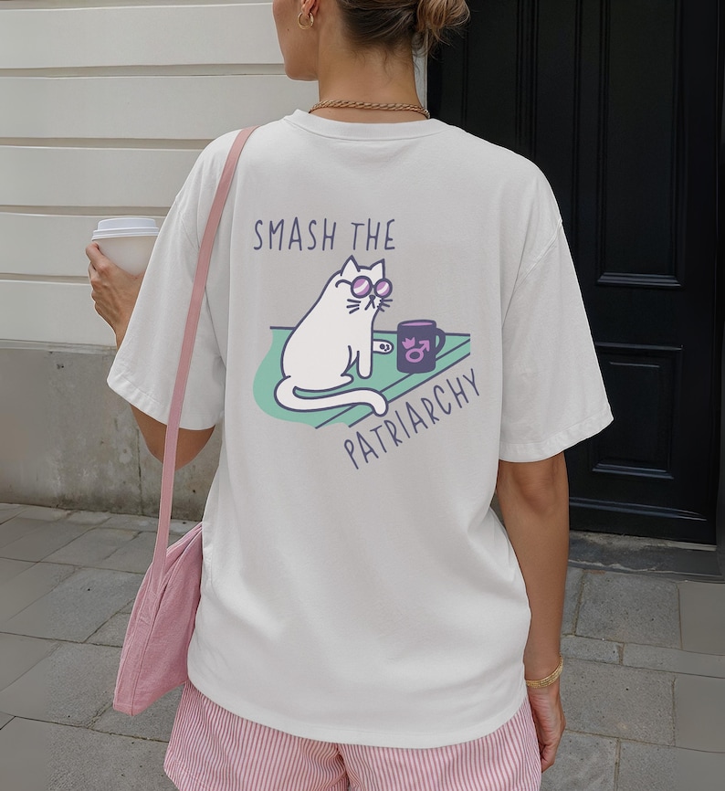 May include: Cream-colored t-shirt with a graphic on the back. The graphic features a white cat wearing sunglasses, a coffee mug, and the text "SMASH THE PATRIARCHY". The cat is sitting on a green surface.