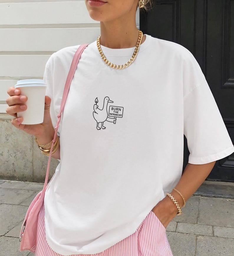 May include: White t-shirt featuring a cartoon duck holding a sign that reads "BURN THE PATRIARCHY". The shirt is paired with pink and white striped pants and a pink purse. The person is also wearing gold jewelry and holding a white coffee cup.