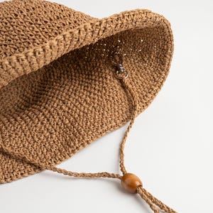 May include: A tan woven bucket hat featuring a crocheted texture and a wide brim. The interior includes a silver-toned metal lobster clasp attached to a thin, braided cord that features a smooth, oval-shaped wooden bead for adjustable tightening.