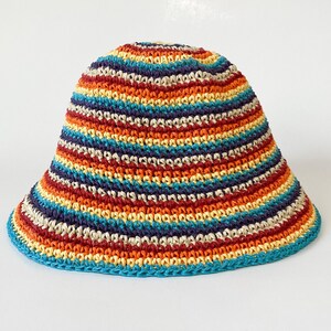 Puede incluir: Un sombrero de cubo de ganchillo a rayas multicolores. El sombrero presenta bandas de rojo, naranja, amarillo, azul y morado. El borde y el borde superior son de color turquesa liso. El sombrero se muestra sobre un fondo blanco.