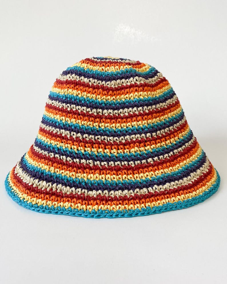 Puede incluir: Un sombrero de cubo de ganchillo a rayas multicolores. El sombrero presenta bandas de rojo, naranja, amarillo, azul y morado. El borde y el borde superior son de color turquesa liso. El sombrero se muestra sobre un fondo blanco.