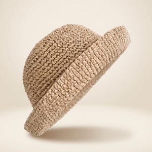 May include: A beige, woven hat with a rounded crown and a short, folded brim. The hat is made of a textured material, likely straw or a similar natural fiber. The hat is angled slightly, showcasing its shape and texture.