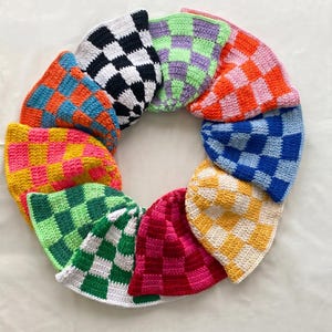 Op de afbeelding: Een verzameling kleurrijke, gehaakte vissershoeden, gerangschikt in een cirkelvormig patroon. Elke hoed heeft een dambordpatroon in verschillende kleurencombinaties, waaronder zwart en wit, oranje en blauw, en roze en geel. De hoeden worden gepresenteerd op een wit oppervlak.