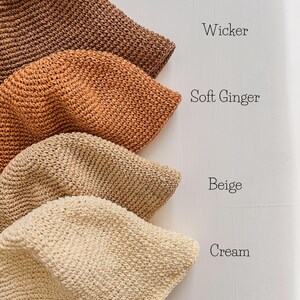 May include: Four crocheted bucket hats in shades of brown, orange, beige, and cream. The hats are displayed in a row, with the names "Wicker", "Soft Ginger", "Beige", and "Cream" written next to each hat. The hats have a textured appearance.