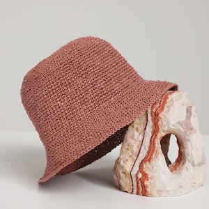 May include: A dusty rose-colored woven bucket hat with a slightly downward-sloping brim. The hat is resting on a textured, light-colored stone with red and brown streaks. The hat's interior is a darker shade of brown.