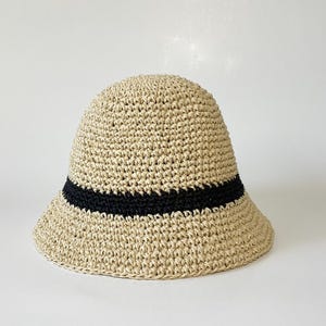 May include: A beige woven bucket hat with a black band. The hat has a rounded crown and a slightly downward-sloping brim. The hat is made of a natural fiber material.