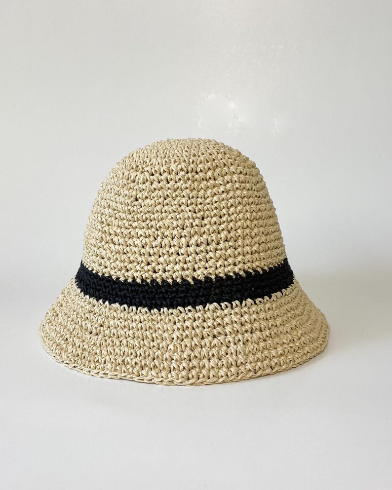 May include: A beige woven bucket hat with a black band. The hat has a rounded crown and a slightly downward-sloping brim. The hat is made of a natural fiber material.
