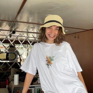 May include: A straw hat with a black band and a white t-shirt with a floral design. The t-shirt is oversized. The person is smiling.