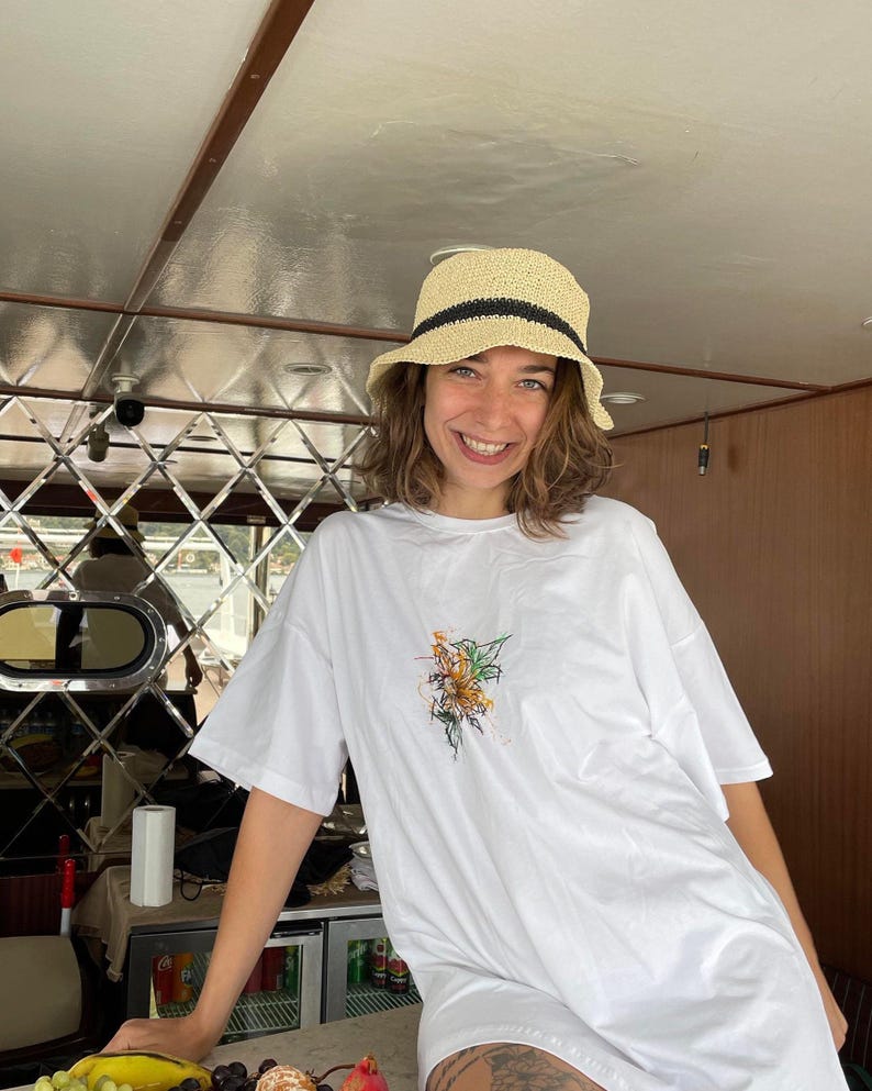 May include: A straw hat with a black band and a white t-shirt with a floral design. The t-shirt is oversized. The person is smiling.