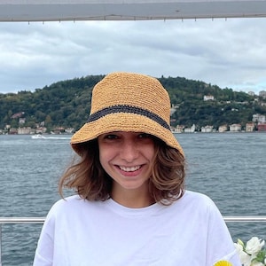 May include: A straw bucket hat with a black band is worn by a person. The hat is a light tan color. The person is wearing a white t-shirt with a graphic design. The background shows a body of water and a green, tree-covered hill.