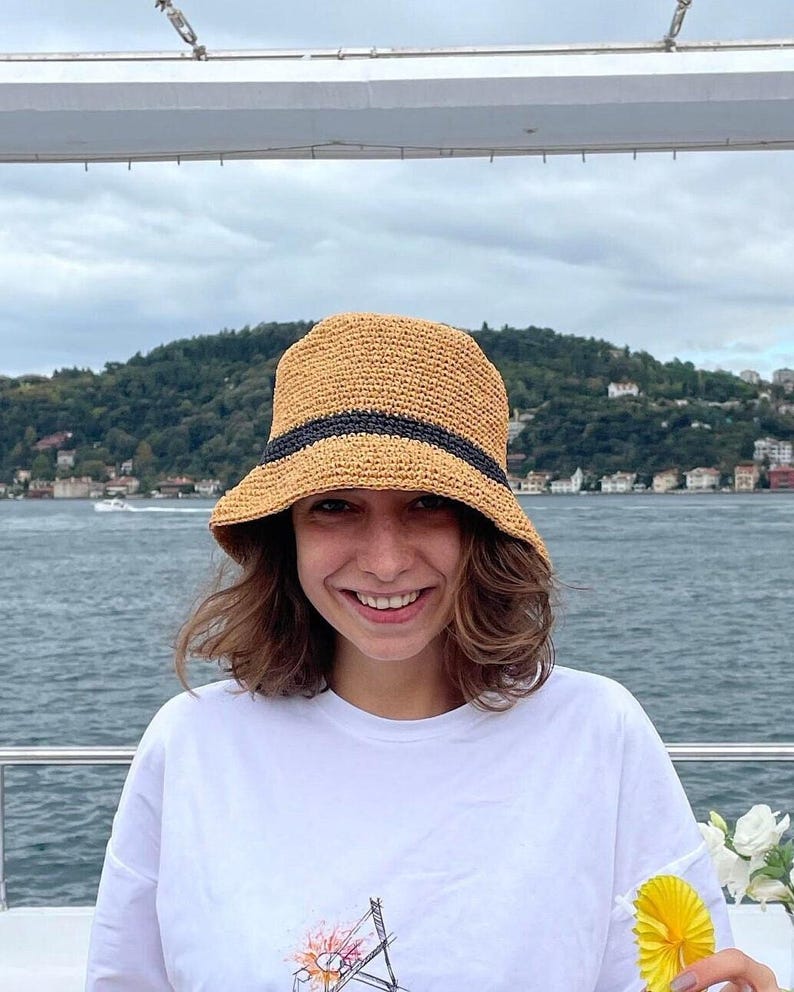 May include: A straw bucket hat with a black band is worn by a person. The hat is a light tan color. The person is wearing a white t-shirt with a graphic design. The background shows a body of water and a green, tree-covered hill.