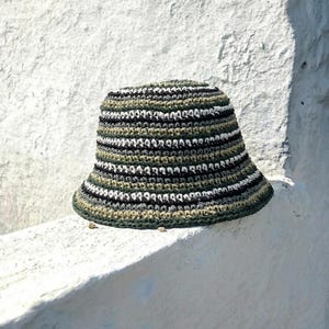 Puede incluir: Sombrero de pescador de ganchillo hecho a mano en tonos verdes, negros, blancos y grises. El sombrero presenta rayas horizontales y un ala ancha. El sombrero est&aacute; sobre una superficie blanca.