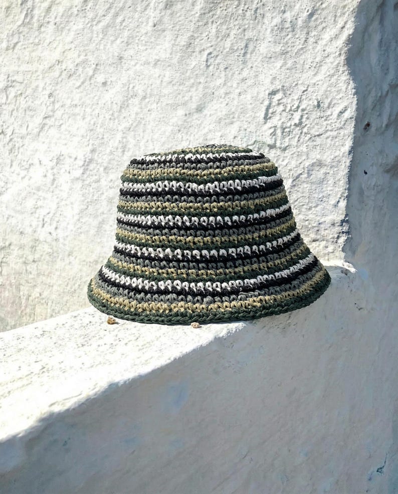 Puede incluir: Sombrero de pescador de ganchillo hecho a mano en tonos verdes, negros, blancos y grises. El sombrero presenta rayas horizontales y un ala ancha. El sombrero est&aacute; sobre una superficie blanca.