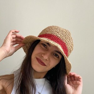 May include: A woven straw hat with a red band. The hat has a rounded crown and a slightly curved brim. The hat is a light tan color with a red band around the crown. The hat is a summer accessory.