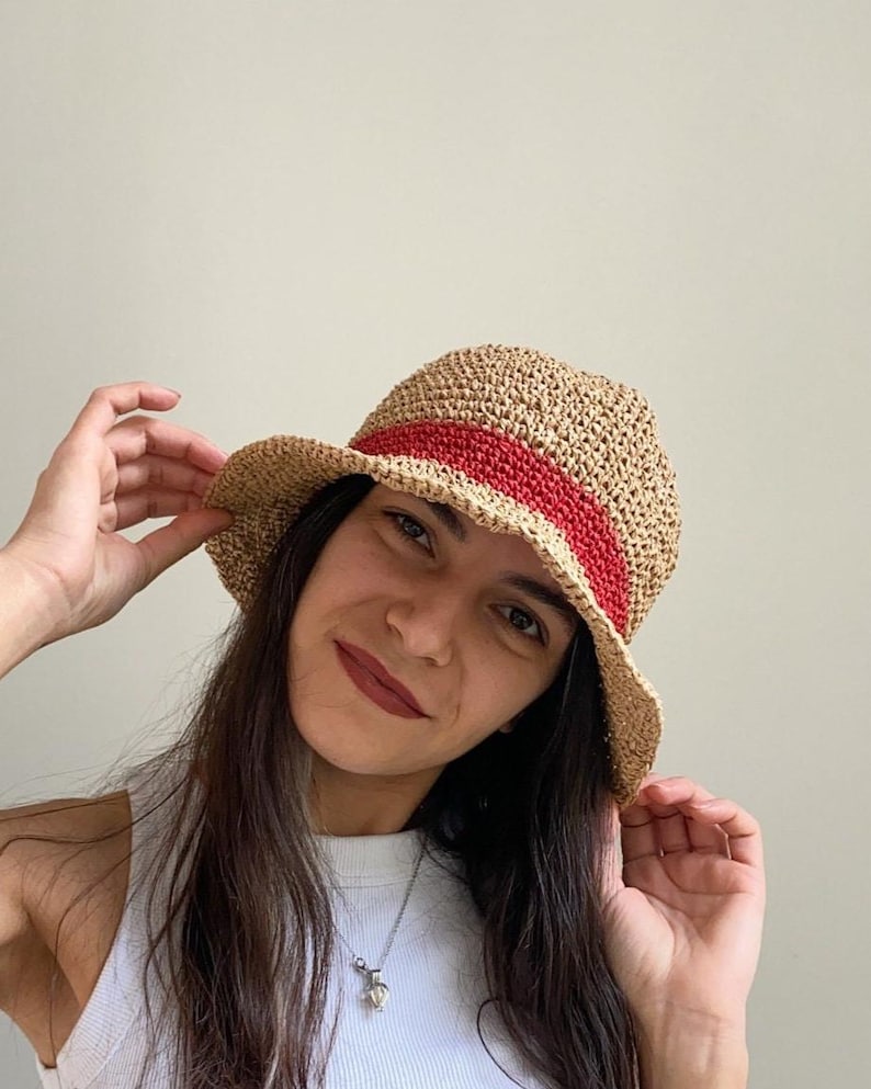 May include: A woven straw hat with a red band. The hat has a rounded crown and a slightly curved brim. The hat is a light tan color with a red band around the crown. The hat is a summer accessory.