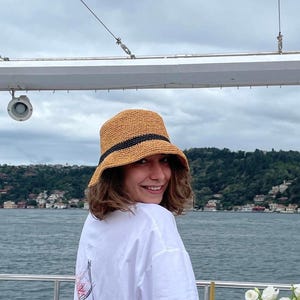 May include: A straw-colored bucket hat with a black band is worn by a person on a boat. The person is wearing a white t-shirt with a graphic design on the back. The background includes water, trees, and a cloudy sky.
