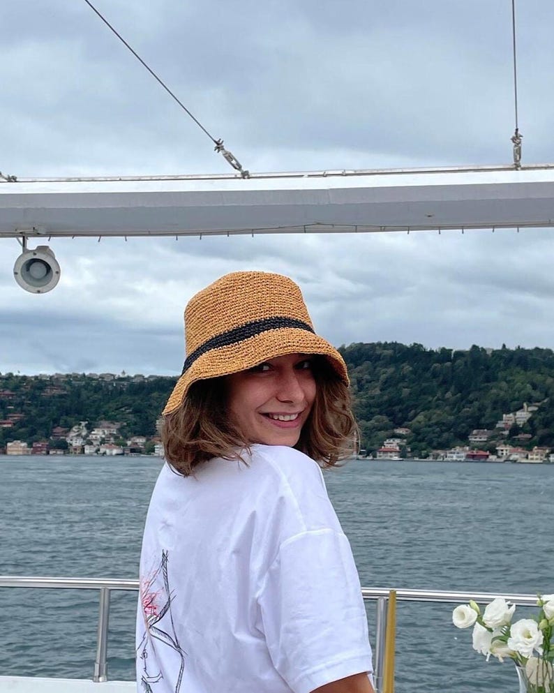 May include: A straw-colored bucket hat with a black band is worn by a person on a boat. The person is wearing a white t-shirt with a graphic design on the back. The background includes water, trees, and a cloudy sky.