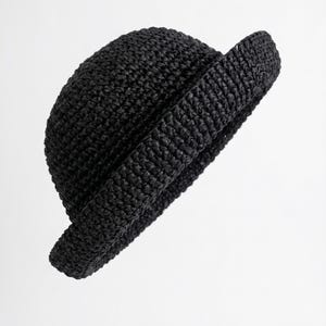 Pode incluir: Um chapéu preto com uma copa arredondada e uma aba curta virada para cima. O chapéu é construído com um material tecido e texturizado com um acabamento escuro consistente, apresentando um padrão denso e entrelaçado de forma estruturada.
