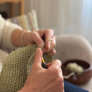 Puede incluir: Primer plano de manos tejiendo con un ganchillo e hilo verde claro. La tela tejida es de color verde claro texturizado. Un cuenco de madera con lana est&aacute; en el fondo. La escena sugiere un proyecto artesanal hecho a mano y acogedor.