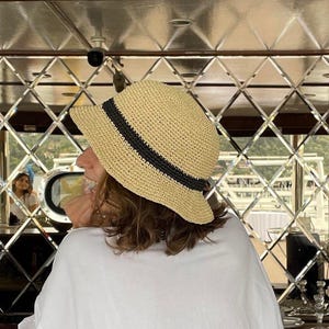 May include: A straw sun hat with a black band is worn by a person. The hat is beige and has a wide brim. The person is wearing a white t-shirt. The background is a mirrored wall.