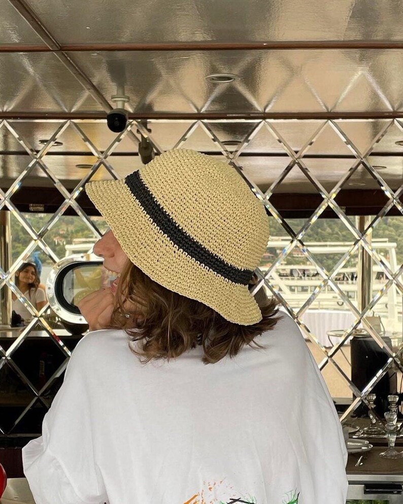 May include: A straw sun hat with a black band is worn by a person. The hat is beige and has a wide brim. The person is wearing a white t-shirt. The background is a mirrored wall.