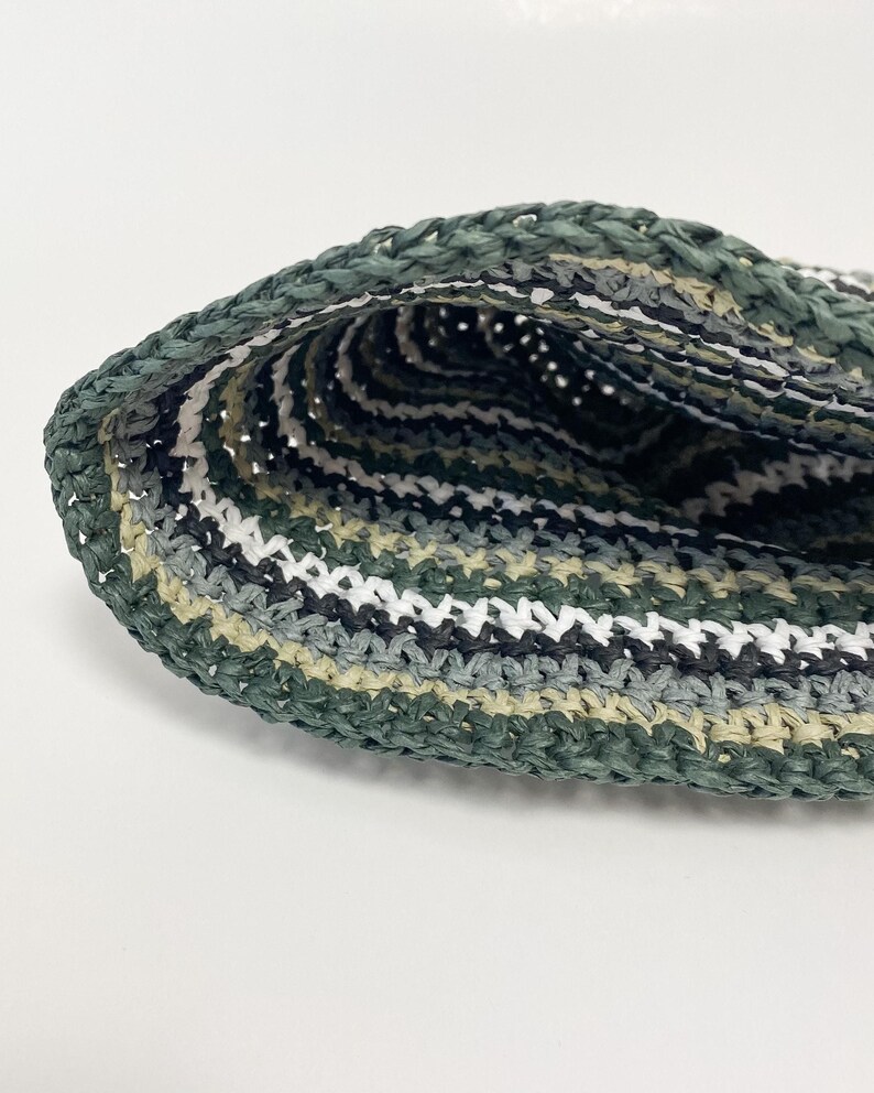 Puede incluir: Primer plano de un sombrero tejido a mano. El sombrero presenta una mezcla de hilos verdes, blancos, grises y negros, creando un patr&oacute;n de rayas. La textura del sombrero es visible, mostrando la artesan&iacute;a. El sombrero es probablemente para protecci&oacute;n solar o moda.