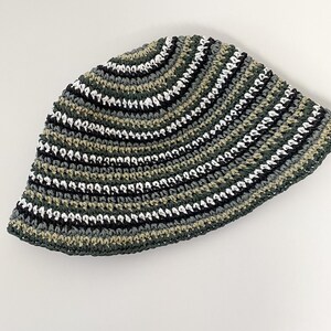 Puede incluir: Un sombrero de pescador de ganchillo con rayas horizontales en tonos de verde, negro, blanco y gris. El sombrero est&aacute; hecho a mano y tiene una corona redondeada y un ala ligeramente inclinada hacia abajo. El patr&oacute;n de color crea una apariencia texturizada.