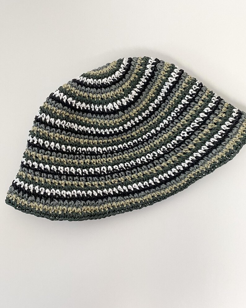 Puede incluir: Un sombrero de pescador de ganchillo con rayas horizontales en tonos de verde, negro, blanco y gris. El sombrero est&aacute; hecho a mano y tiene una corona redondeada y un ala ligeramente inclinada hacia abajo. El patr&oacute;n de color crea una apariencia texturizada.