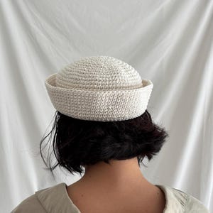 May include: A cream-coloured, crocheted hat with a rounded crown and a slightly upturned brim. The hat is made of a textured yarn and appears to be handmade. The hat is a stylish accessory.