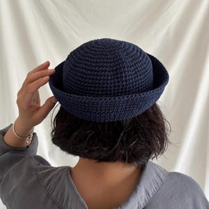 Puede incluir: Un sombrero de ganchillo azul marino con una corona redondeada y un ala ancha. El sombrero está hecho a mano y parece estar hecho de un hilo suave. El sombrero está siendo sostenido por una persona.