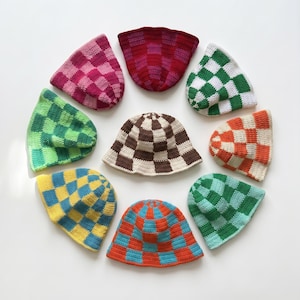 Op de afbeelding: Een verzameling van tien gehaakte bucket hats met een dambordpatroon in verschillende kleuren, waaronder roze, rood, groen, wit, oranje, blauw, geel en bruin. De hoeden zijn in een cirkel gerangschikt op een witte achtergrond.
