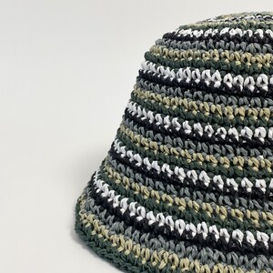 Puede incluir: Un sombrero de pescador tejido a ganchillo con un dise&ntilde;o de camuflaje. El sombrero presenta rayas horizontales verdes, negras, blancas y grises. El borde es de color verde oscuro liso. El sombrero est&aacute; hecho de un material texturizado.