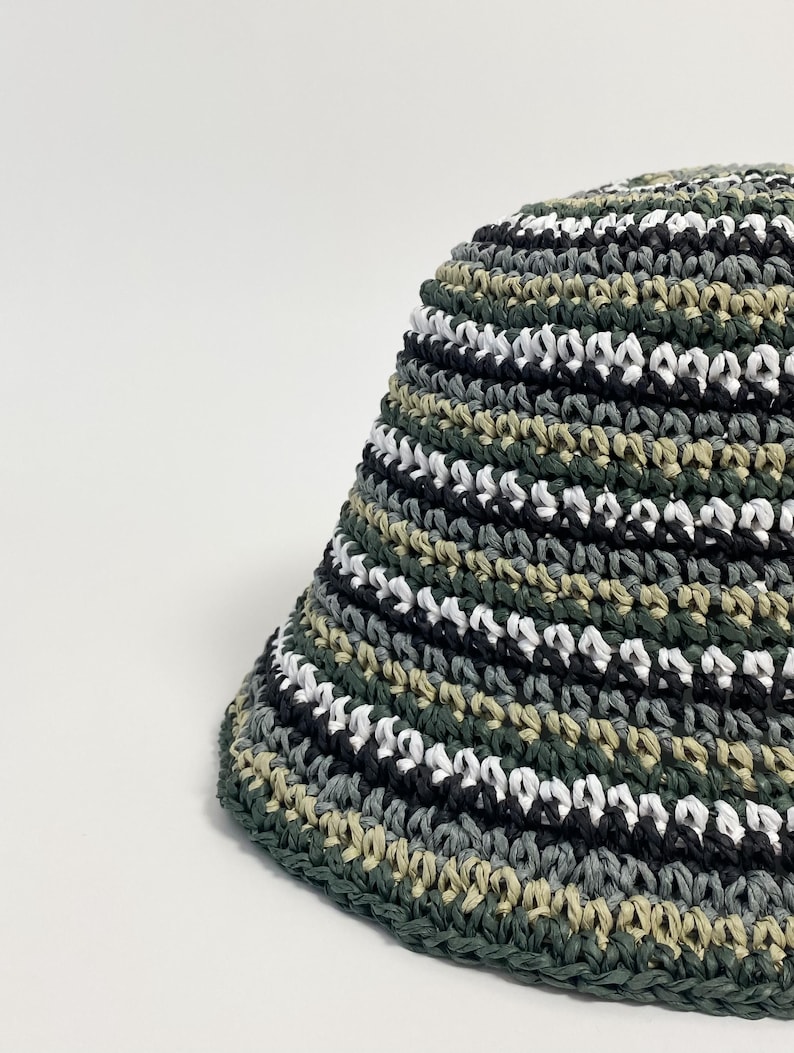 Puede incluir: Un sombrero de pescador tejido a ganchillo con un dise&ntilde;o de camuflaje. El sombrero presenta rayas horizontales verdes, negras, blancas y grises. El borde es de color verde oscuro liso. El sombrero est&aacute; hecho de un material texturizado.
