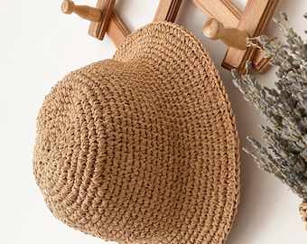 May include: A woven straw hat with a wide brim hangs on a wooden wall rack. The hat is a light brown color and has a textured appearance. The rack is made of light-colored wood and has several pegs for hanging items. A bundle of dried lavender is visible.