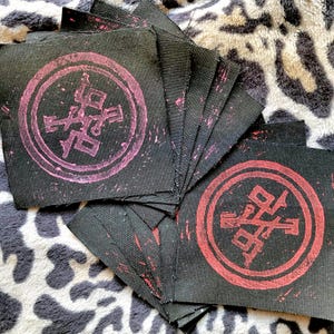 Può includere: Un mucchio di toppe di stoffa nera con un simbolo rosa e rosso stampato su di esse. Il simbolo è un cerchio con una croce all'interno, e le lettere "SO" sono visibili al centro della croce.