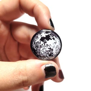 May include: A black and white circular button featuring a detailed illustration of the moon. The button has a glossy finish and is held between fingers against a white background. The design shows the moon's surface.