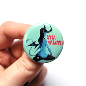 Op de afbeelding: Een tealblauwe knop met een cartoon afbeelding van een vrouw met blauwe huid, hoorns en een staart. De vrouw draagt een zwarte kousenband en kousen. De tekst "STAY WICKED!" is in roze op de knop gedrukt.