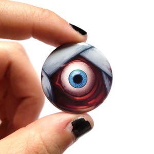 Könnte beinhalten: Ein runder Button mit einer detaillierten Illustration eines blauen Auges mit roten und weißen Akzenten. Das Auge ist der Mittelpunkt, umgeben von einem roten und weißen Hintergrund. Der Button wird in einer Hand gehalten.